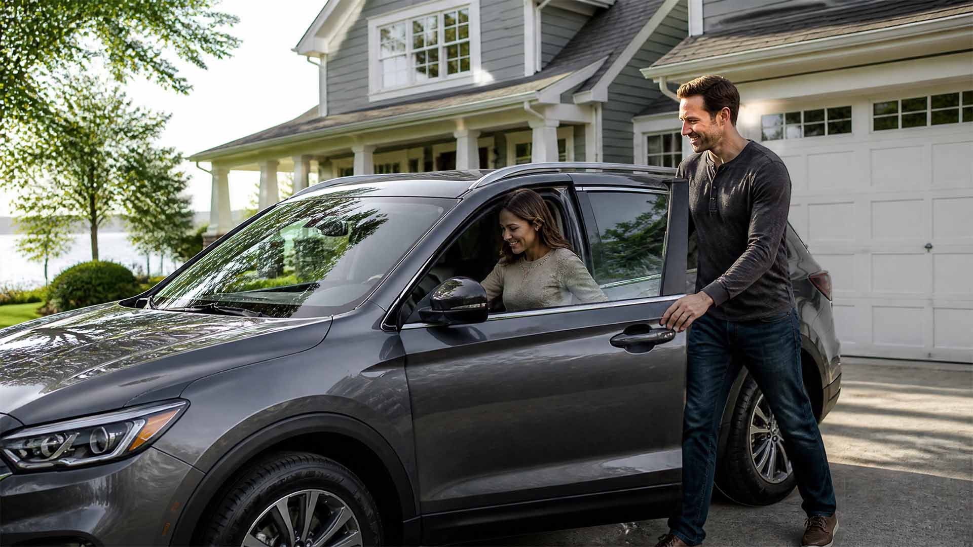 A smiling man opens the passenger door of a grey crossover SUV parked in a driveway for a woman sitting inside, with a two-story house in the background - www.johnscottinsurance.com